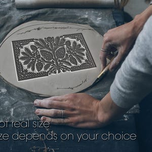 May include: A flat, circular piece of light grey clay featuring a rectangular stamped design of a central tree with pinecones and foliage, bordered by intricate patterns. The text "Not real size. Size depends on your choice" is visible.