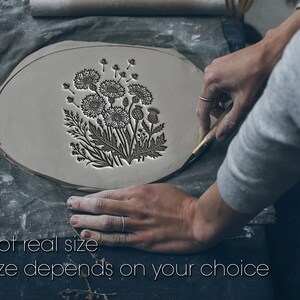 Può includere: Un pezzo di argilla grigia, piatto e di forma ovale, con una dettagliata illustrazione botanica in rilievo di fiori e foglie di cardo. Il testo "Not real size" e "Size depends on your choice" è stampato in bianco in fondo all'immagine.