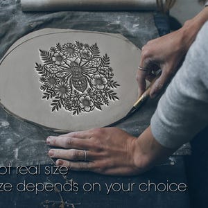Può includere: Una lastra di argilla grigia con un design profondamente impresso di un'ape centrale circondata da vari fiori e foglie di felce. Il testo Not real size Size depends on your choice è stampato in bianco nella parte inferiore.