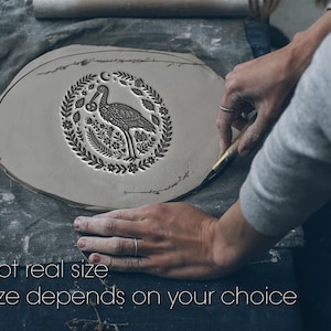 Op de afbeelding: Een plat, cirkelvormig stuk grijze klei met een gestempeld ontwerp van een ooievaar die een krakeling vasthoudt, omringd door een krans van bladeren, bloemen en een maansikkel. De tekst "Not real size. Size depends on your choice" is zichtbaar.