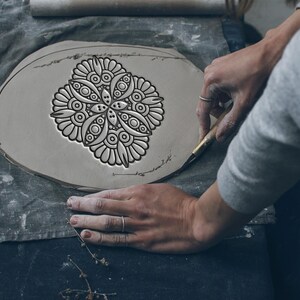 Puede incluir: Manos trabajando en una pieza de arcilla con un diseño floral decorativo. La arcilla es de color gris claro y el diseño es de un gris más oscuro. Se utiliza una herramienta para cortar alrededor de la pieza de arcilla. El fondo incluye un rodillo y un paño gris.