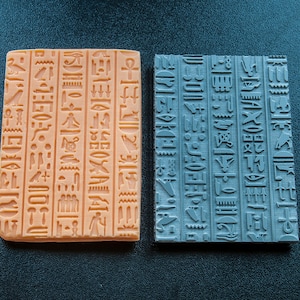 May include: Two rectangular blocks with ancient Egyptian hieroglyphs. One block is a light orange color, and the other is a muted teal. The hieroglyphs are raised and detailed, showcasing various symbols and characters. The blocks are placed on a dark surface.