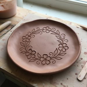 Può includere: Un piatto di argilla non cotta con un motivo floreale. Il piatto è di colore terracotta naturale, con un motivo a ghirlanda floreale intagliato sul bordo. Il piatto è rotondo e poggia su una superficie di legno. Diametro di circa 20 cm.