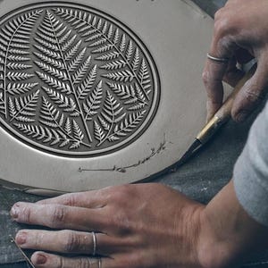 Peut inclure: Une assiette en céramique avec un motif de fougère détaillé sculpté à sa surface. La personne utilise un outil de sculpture. Le motif de fougère est de forme circulaire au centre de l'assiette.