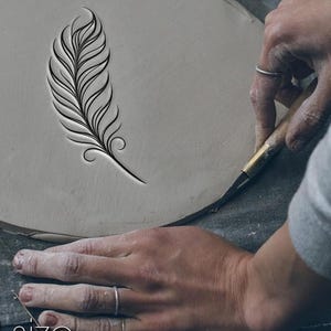 May include: A detailed feather design is carved into a light gray clay surface. The image shows the carving process, with hands holding a tool to create the design. The feather has intricate lines and curves.