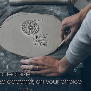 May include: A gray, oval-shaped clay piece with a dandelion design and the text "Words are only Duff". The clay is being shaped with a tool. The text "Not real size" and "Size depends on your choice" are also visible.