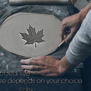 May include: A detailed maple leaf design carved into a light grey clay surface. The text "Not real size, Size depends on your choice" is visible. The image shows the clay being worked on a grey cloth surface.