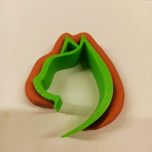 Peut inclure: Un emporte-pièce vert en forme de chat est posé sur une base brun-rouge. Le découpoir représente le profil d'une tête de chat, avec des oreilles pointues et un museau incurvé. Le plastique est texturé.