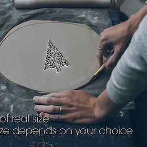 Puede incluir: Una pieza de arcilla ovalada de color gris claro con un diseño de árbol de Navidad recortado. El árbol está relleno con un patrón de hojas negras. El texto "Not real size" y "Size depends on your choice" está en la parte inferior.