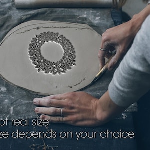 Op de afbeelding: Een grijze kleiplaat met een cirkelvormig reliëfontwerp van een krans bestaande uit dennenappels, hulstbladeren en kleine sterren. De tekst "Not real size" en "Size depends on your choice" staat in het wit onderaan de afbeelding.
