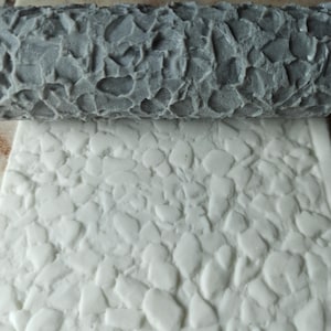 May include: A gray textured rolling pin with a pattern of small, irregular shapes. The rolling pin is used to create a stone-like texture on a white surface.