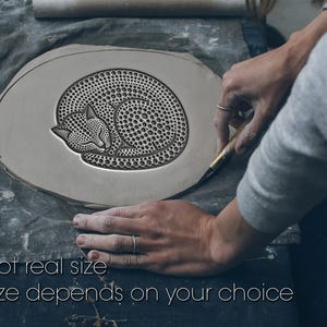 Può includere: Un'opera d'arte in argilla con un disegno di gatto che dorme. Il gatto è rannicchiato, con un motivo dettagliato di punti che copre il suo corpo. Il testo "Not real size" e "Size depends on your choice" è in basso nell'immagine.