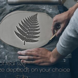 Può includere: Un disegno di foglia di felce è scolpito su una superficie di argilla di colore chiaro. Il testo "Not real size" e "Size depends on your choice" è visibile. L'artigiano sta usando uno strumento per rifinire i bordi dell'argilla.