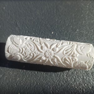 Può includere: Un mattarello cilindrico bianco caratterizzato da un intricato motivo floreale profondamente impresso su tutta la superficie. Il design è composto da vari fiori stilizzati, foglie e motivi a viticcio che creano un effetto in rilievo.