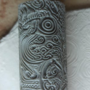 Peut inclure: Objet cylindrique gris avec des motifs complexes et tourbillonnants. Le design présente des formes et des lignes stylisées répétitives, possiblement sculptées ou en relief. La surface est d'un gris discret, avec un design détaillé.