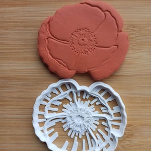 Può includere: Un tagliabiscotti e un timbro a forma di papavero sono esposti su una superficie di legno. Il timbro è di colore arancione-rossastro e il tagliabiscotti è bianco. Il design del papavero è dettagliato, mostrando petali e il centro del fiore.