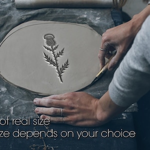 Könnte beinhalten: Ein Disteldesign ist in ein ovales Tonstück geschnitzt. Der Text "Not real size" und "Size depends on your choice" befindet sich unten. Das Bild zeigt den Prozess der Töpferei.