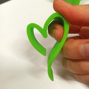 Op de afbeelding: Een felgroen hartvormig object, mogelijk een koekjesvorm of knutselgereedschap, wordt door een persoon vastgehouden. Het object heeft een glad, gebogen ontwerp en een kleine opening aan de bovenkant. De kleur is een levendig, opvallend groen.