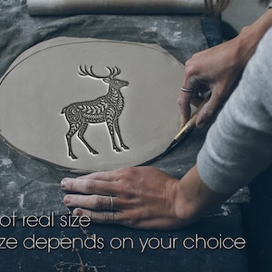 Peut inclure: Une pièce d'argile gris clair avec un motif de cerf détaillé. Le cerf est gravé de motifs complexes. Le texte "Not real size" et "Size depends on your choice" est en bas. L'image montre le cerf en cours de sculpture.