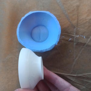 May include: A light blue silicone mould with a circular opening and a smooth interior, paired with a white, conical, 3D-printed object featuring a subtle, fine-lined, diagonal texture on its exterior surface, shown from a top-down perspective.