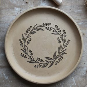 Op de afbeelding: Een beige keramische plaat met een donkerbruin bloemenkransontwerp. De krans is samengesteld uit bladeren en kleine bloemclusters. De plaat heeft een licht verhoogde rand en een glad oppervlak. De plaat is waarschijnlijk handgemaakt.