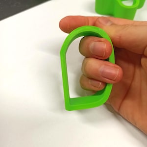 Puede incluir: Un objeto verde brillante, arqueado, impreso en 3D, posiblemente un asa o parte de un objeto más grande. El objeto se sostiene en una mano, mostrando su forma y color sobre un fondo blanco. El diseño es simple y moderno.