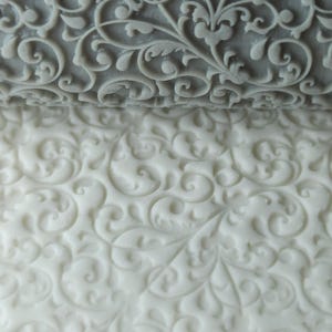May include: A close-up of a gray rolling pin with a decorative floral pattern. The rolling pin is used to create an embossed pattern on fondant or dough.