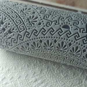 May include: A gray rolling pin with a detailed embossed pattern of Greek key and floral designs. The rolling pin is used to create decorative patterns on fondant or dough.