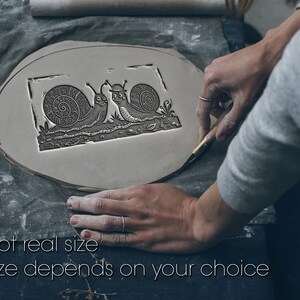 Könnte beinhalten: Ein flaches, kreisförmiges Stück grauer Ton mit einem geprägten rechteckigen Design von zwei Schnecken mit detaillierten Gehäusemustern. Eine Schnecke trägt einen kleinen Hut. Der Text "Not real size. Size depends on your choice" ist unten aufgedruckt.