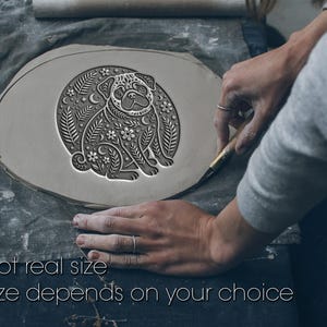 May include: A gray clay pug design with floral and crescent moon accents. The text "Not real size. Size depends on your choice" is visible. The pug is surrounded by a circular design. The image shows the clay being worked on.