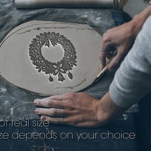 Op de afbeelding: Een grijze kleiplaat met een diep reliëf van een kerstkrans met een centrale strik, hulstbladeren, bessen, sterren, een bel en ornamenten. De tekst "Not real size. Size depends on your choice" staat onderaan de afbeelding weergegeven.