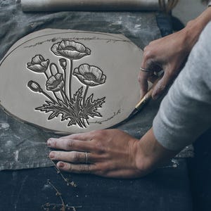 May include: A person's hands are shown carving a poppy flower design into a gray clay plate. The plate is oval-shaped and rests on a gray cloth. The poppy design is detailed with black lines.