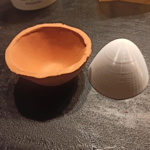 Op de afbeelding: Een open kom van terracotta kleur met een ruwe rand en een gladde binnenkant, naast een wit, kegelvormig object. De kegel heeft een geribbelde oppervlakte, met concentrische cirkels aan de bovenkant.