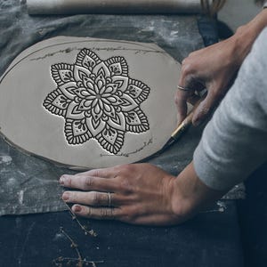 May include: A detailed, black mandala design is carved into a gray clay disc. The artisan's hands, wearing a ring, carefully work with a tool to refine the edges of the design. The clay sits on a gray cloth, with a rolling pin visible in the background.