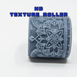 Könnte beinhalten: Eine graue HD-Texturrolle mit floralem Design. Die Rolle weist ein detailliertes Muster einer zentralen Blume auf, die von komplizierten Blatt- und Punktmustern umgeben ist. Der Text "HD TEXTURE ROLLER" ist in Blau und Weiß dargestellt.