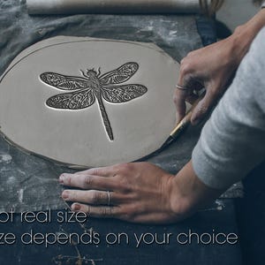 May include: A flat, circular piece of grey clay featuring a detailed, embossed dragonfly design with intricate wing patterns. The clay is being shaped with a metal tool. The text "Not real size. Size depends on your choice" is visible.