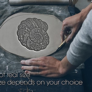 Op de afbeelding: Een plat, ovaal stuk grijze klei met een diep reliëf van een nautilusschelp, gevuld met ingewikkelde bloem- en bladpatronen. De tekst "Not real size. Size depends on your choice" is onderaan in het wit afgedrukt.
