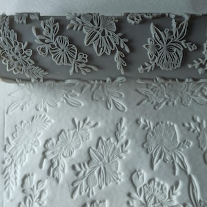 Op de afbeelding: Een grijze deegroller met een bloemenpatroon. De deegroller wordt gebruikt om een ​​reliëfpatroon op fondant of deeg te creëren. Het ontwerp bevat delicate bloemen en bladeren.
