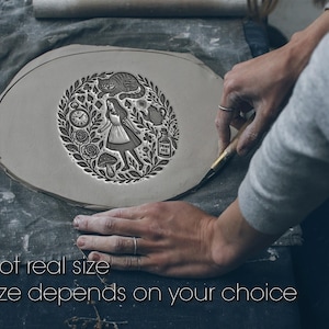 May include: A circular clay stamp featuring an intricate illustration of a girl, a cat, a pocket watch, a teapot, a mushroom, and a bottle labeled "DRINK ME" surrounded by foliage. The text "Not real size. Size depends on your choice" is visible.