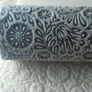 Può includere: Un mattarello grigio con un motivo floreale. Il motivo è in rilievo sul mattarello e presenta un motivo ripetitivo di foglie e fiori.