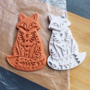 May include: Two fox-shaped cookie cutters. One is orange, the other is white. Both feature detailed floral and celestial designs. The orange cutter is a finished cookie, while the white one is the cutter itself. They are on a wooden surface.