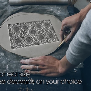 May include: A gray, oval-shaped piece of clay with a rectangular floral pattern carved into its center. The text "Not real size" and "Size depends on your choice" is at the bottom. The image shows hands working on the clay.