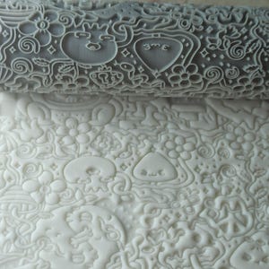 May include: A gray rolling pin with a raised design of various doodles, including hearts, stars, and swirls. The rolling pin is being used to create an impression in white dough.
