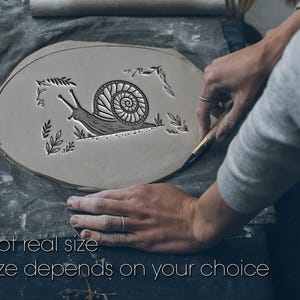 Op de afbeelding: Een plat, ovaal stuk grijze klei met een reliëf van een slak omringd door kleine bladmotieven. De tekst "Not real size" en "Size depends on your choice" staat in het wit onderaan de afbeelding gedrukt.
