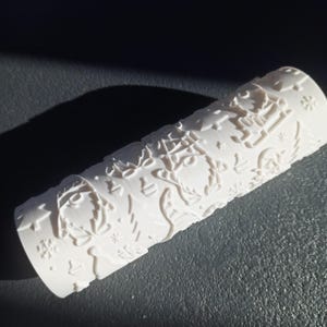 Op de afbeelding: Een witte cilindrische deegroller met een herhalend reliëfpatroon van vakantie-iconen, waaronder kabouters, sneeuwvlokken, kerstbomen en ornamenten. Het oppervlak is voorzien van deze verhoogde ontwerpen voor het bedrukken van deeg.