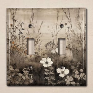 May include: A double light switch cover with a floral design. The cover features a watercolor-style illustration of white flowers and foliage in shades of brown and gray against a light brown background.