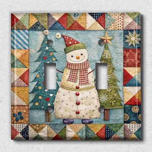 May include: A light blue and white light switch cover with a patchwork design featuring a snowman and two Christmas trees. The snowman is wearing a red and white hat and scarf. The trees are green with white snowflakes.