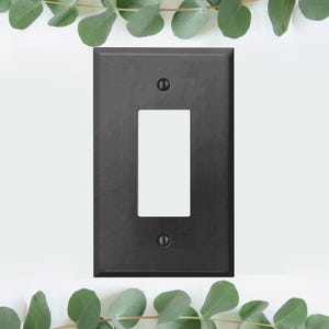 May include: A black metal light switch plate with a single switch opening. The plate has a matte finish and is surrounded by a white background.