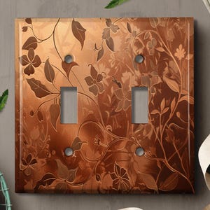 May include: A copper-colored light switch cover with a floral pattern. The switch cover has two toggle switches.