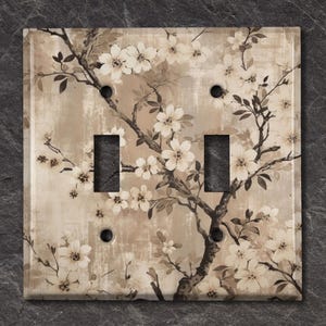 May include: A double light switch cover with a floral design. The cover features a beige background with a brown tree branch and white flowers.